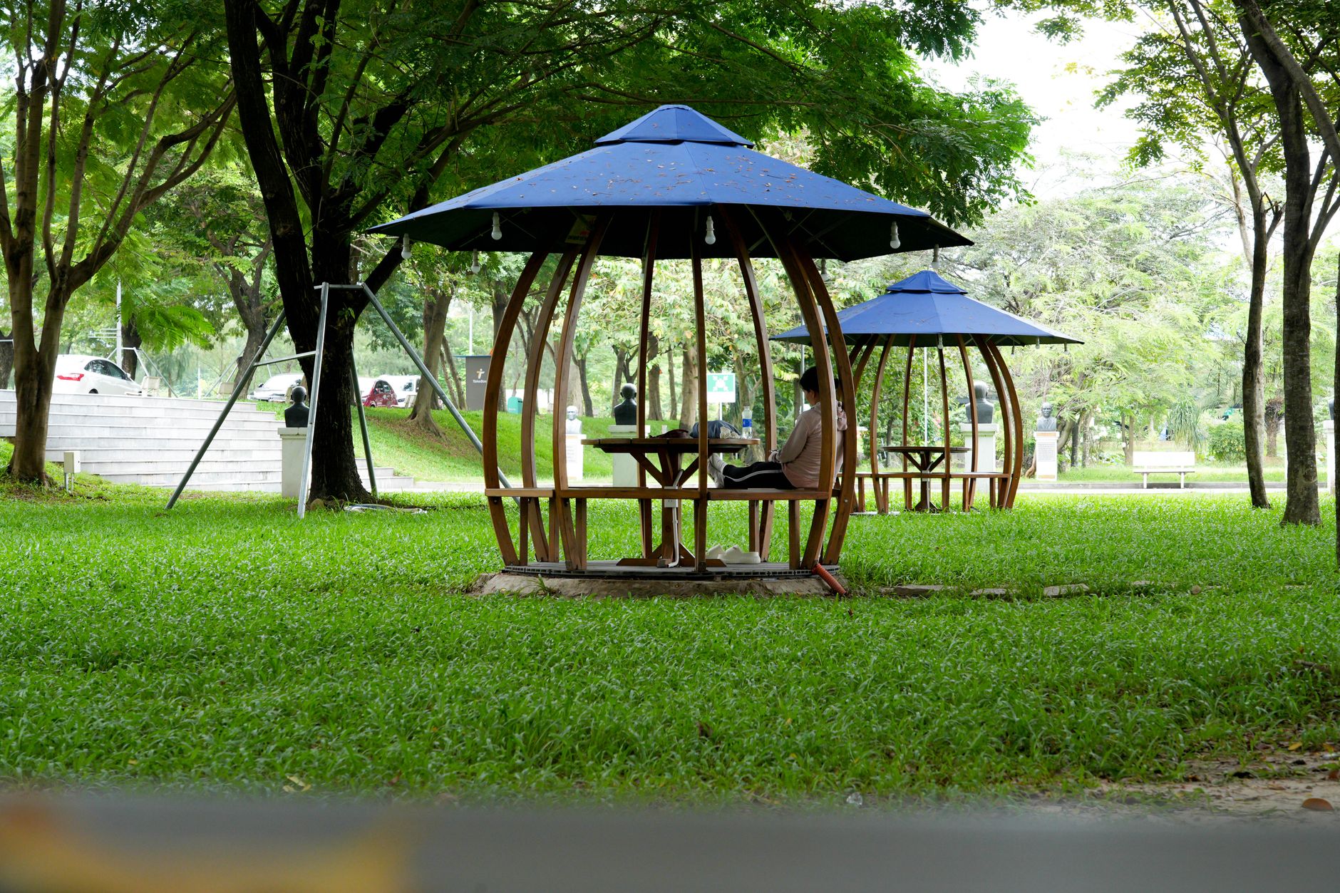 Desain paviliun sederhana dengan atap kaca, struktur kayu ringan, dan taman tropis di sekitarnya.