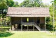 Rumah sederhana di kampung dengan tiga kamar tidur, atap genteng, dan taman kecil.