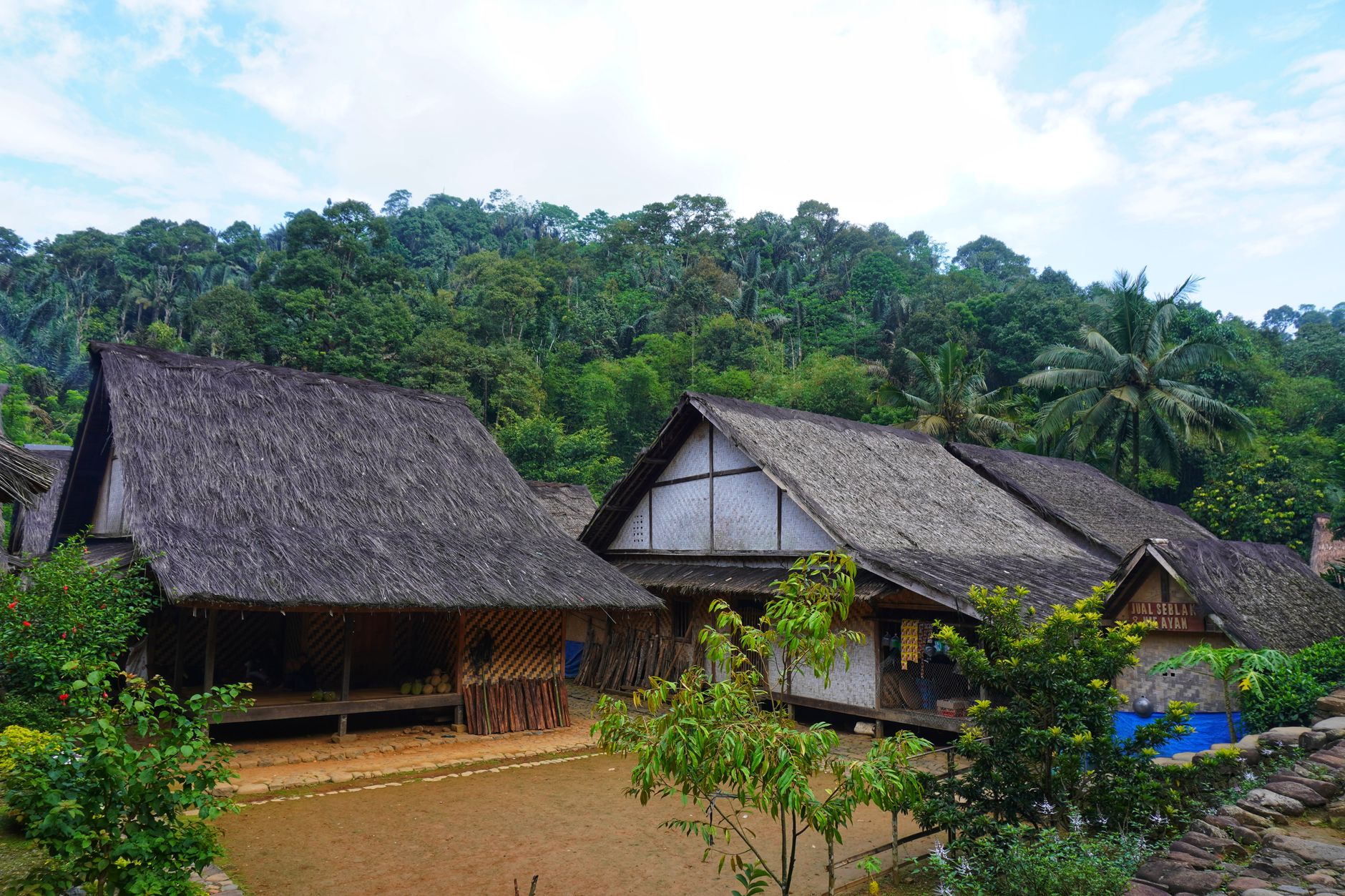 Rumah desa sederhana dengan atap genteng, dinding kayu, dan taman kecil di depan.